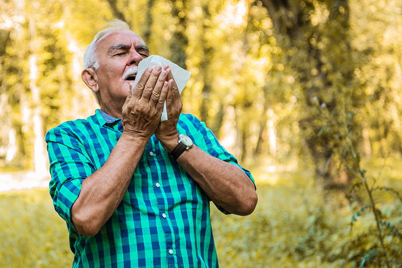 Man with graying hair standing in a park or wooded area, preparing to sneeze
